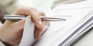 Close-up of hand holding pen with paper over pile of documents