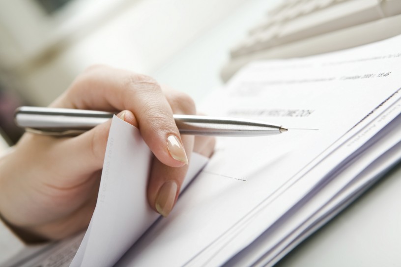 Close-up of  hand holding pen with paper over pile of documents